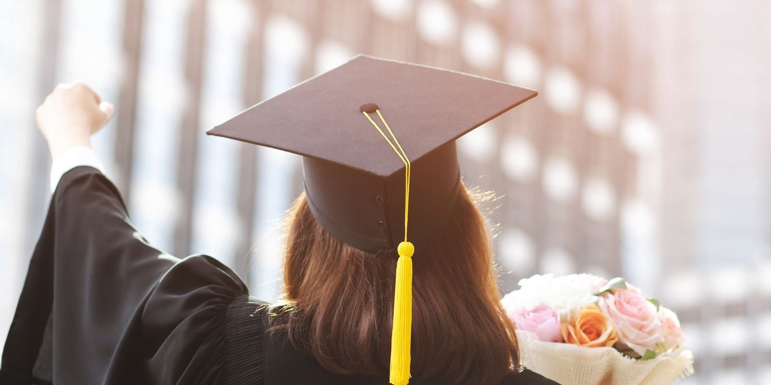 back shot of a person wearing a black graduation cap and gold while holding a fist in the air, triumphantly