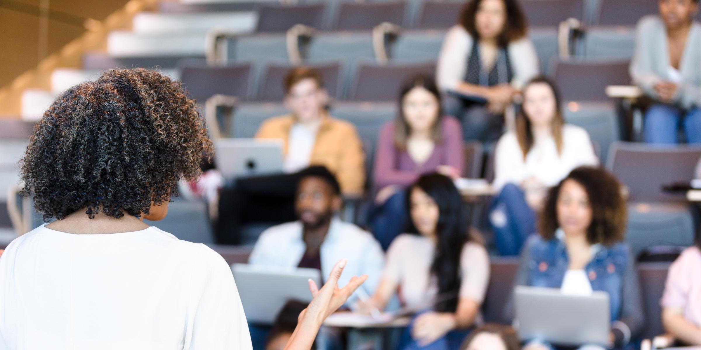 a professor with their back to the camera facing students in a classroom in a blurred background