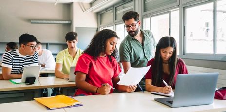 a group of students at two tables looking at papers and laptops inside a classroom